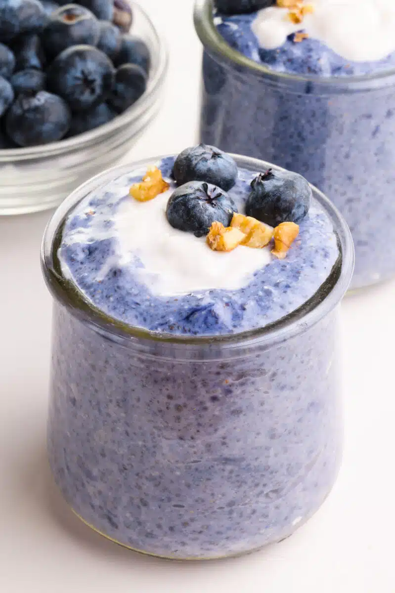 Two glass jars have blue chia pudding in them. They are topped with vegan yogurt, chopped walnuts, and fresh blueberries. There is a bowl with more blueberries in the background.