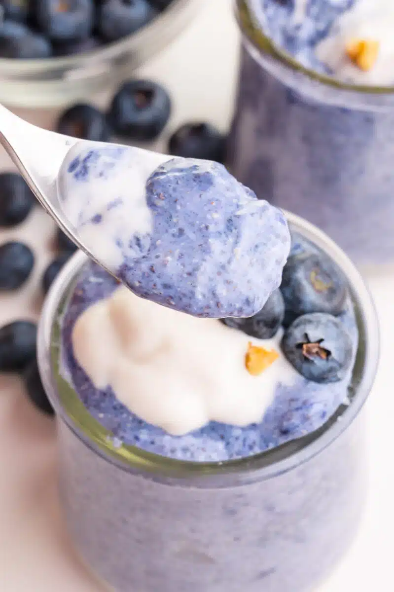 A spoonful of blue chia pudding hovers over the rest of the dish. There is another dish and fresh blueberries in the background.