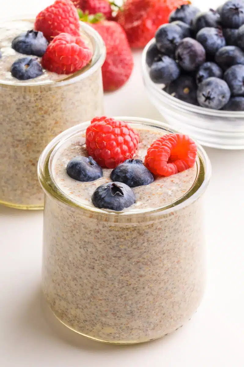 A glass dish holds chia flax pudding with blueberries and raspberries on top. It sits in front of another bowl of the pudding and fresh berries.