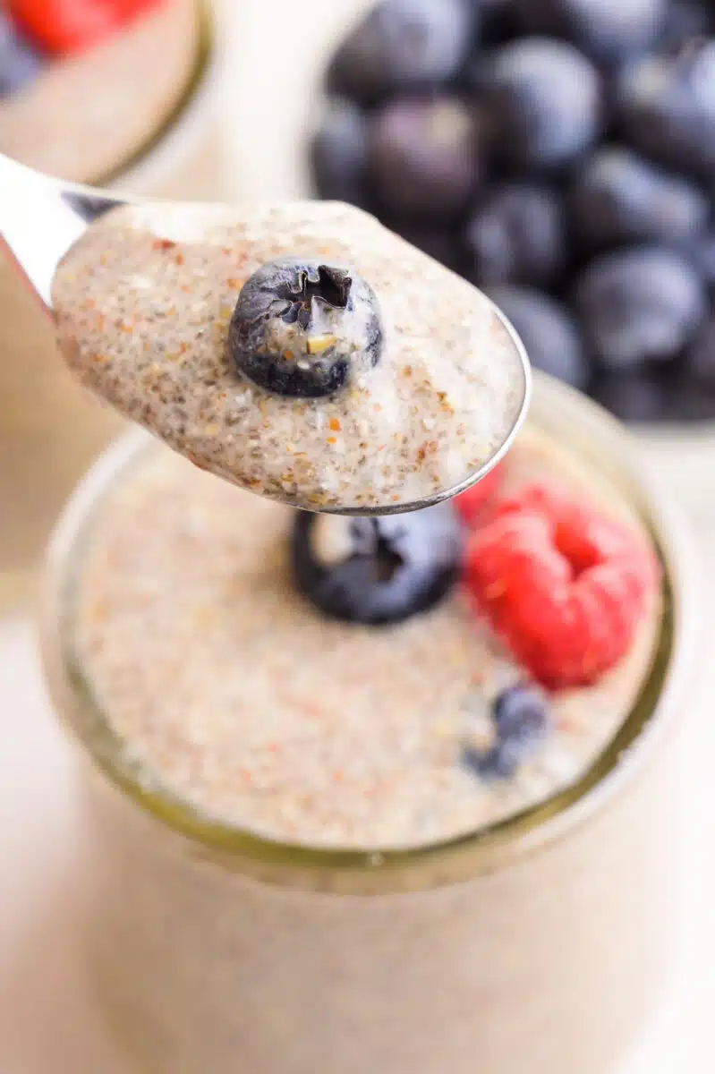 A spoonful of chia pudding has a blueberry in it, hovering over the rest of the bowl. There are fresh blueberries in the background.