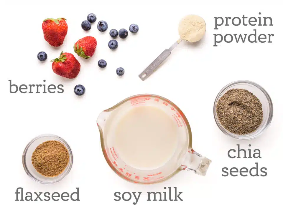 Ingredients are laid out on a table. The labels next to them read, protein powder, chia seeds, soy milk, flaxseed, and berries.