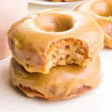 A closeup of a stack of vegan maple donuts. the top one has a bite taken out. There is another donut and a coffee mug visible in the background.