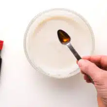 A hand holds a measuring spoon with maple extract over a small bowl with icing in it. There is a bottle of maple extract beside the bowl.