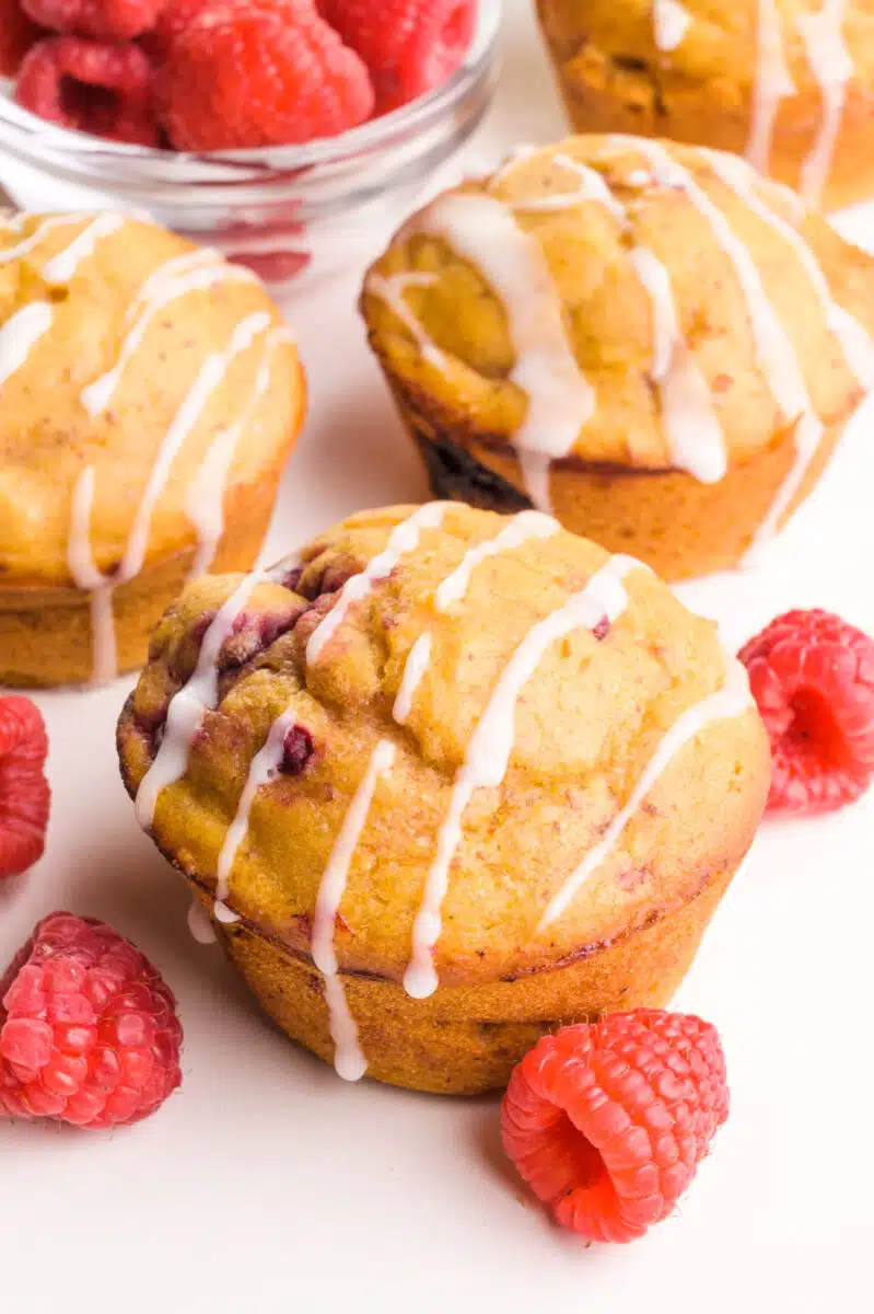 Several muffins sit next to fresh raspberries. The muffins have drizzles of icing on top.