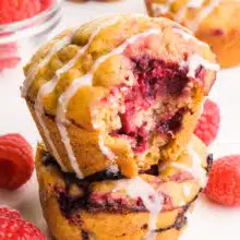 A closeup of vegan raspberry muffins in a stack. The top one has a bite taken out. It sits beside fresh raspberries and muffins.