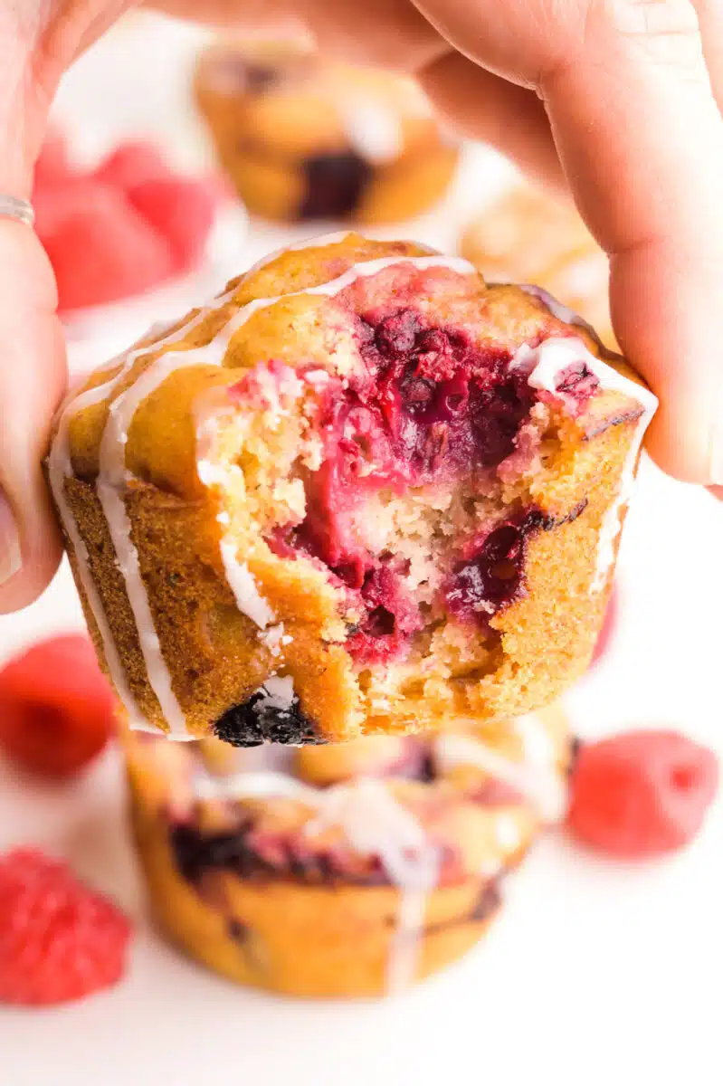A hand holds a vegan raspberry muffin with a bite taken out, revealing lots of raspberries inside.