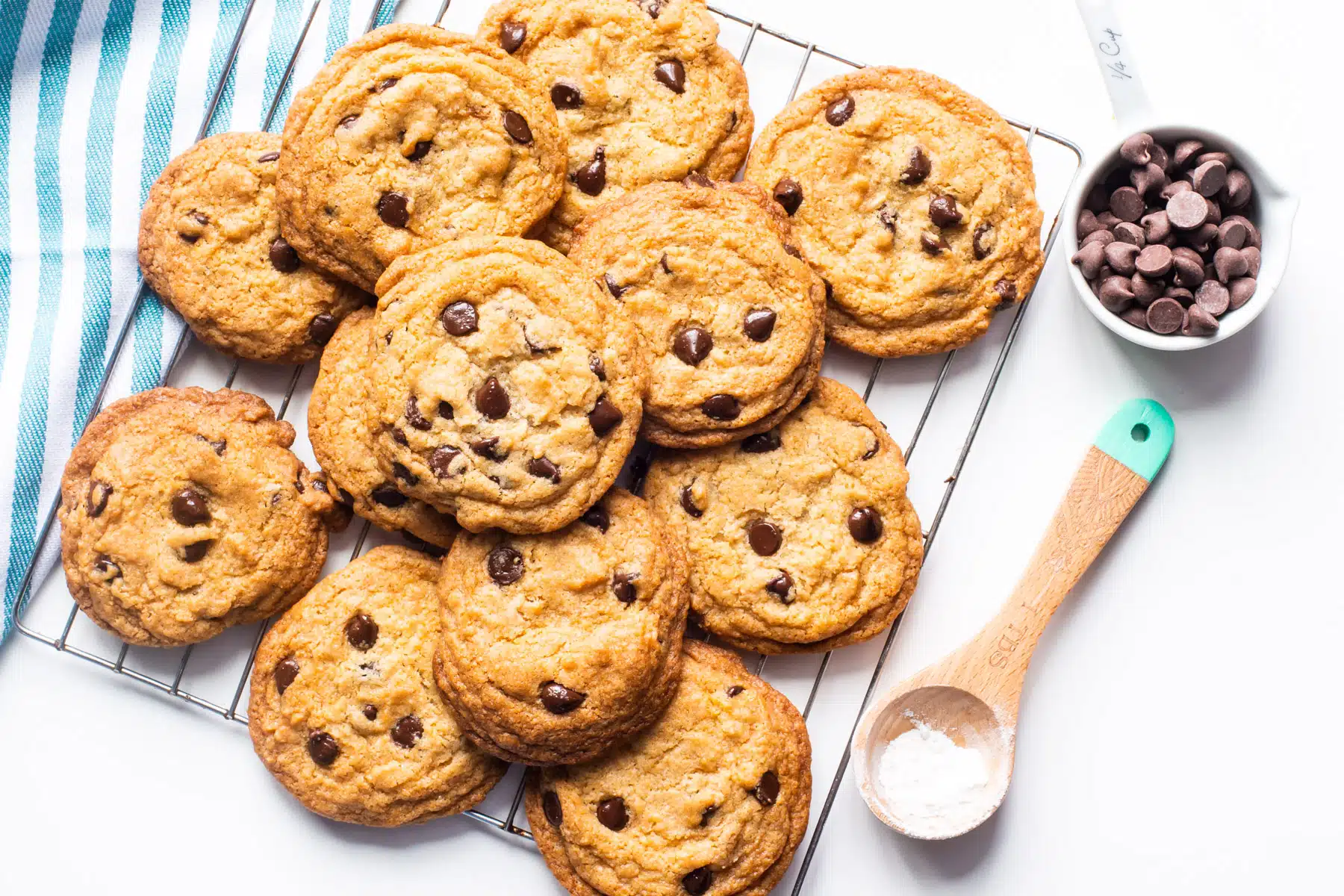 Freshly baked chocolate chip cookies are cooling on a wire rack. A cup of chocolate chips sits beside them along with a measuring spoon with baking soda in it.