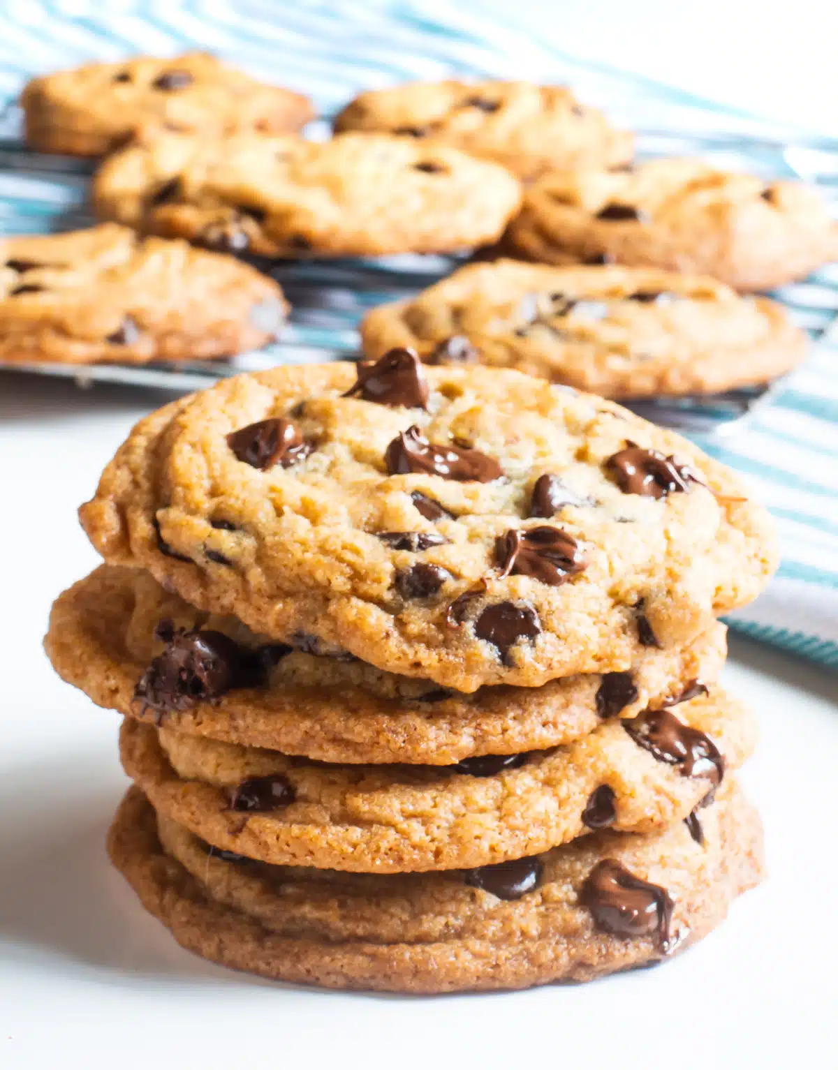 A close-up on a stack of four chocolate chip cookies sitting on on top of the other. More freshly baked chocolate chip cookies are in the background.