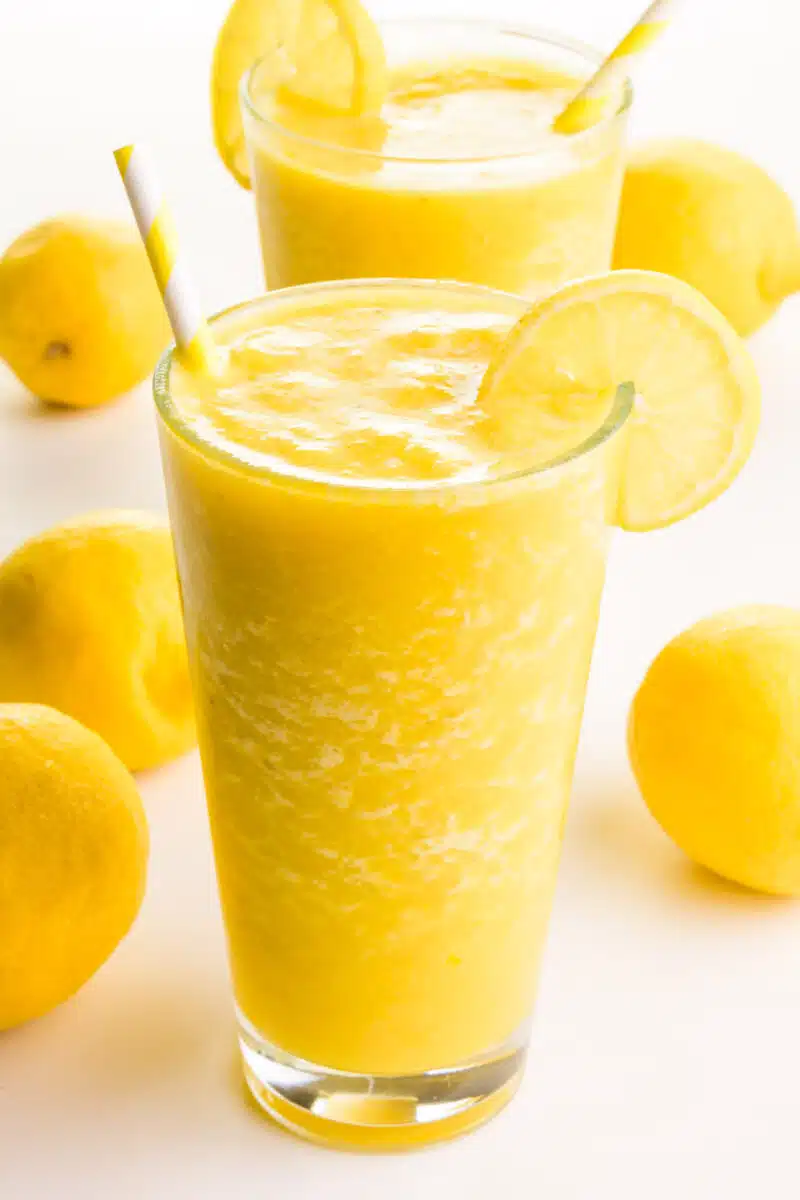 Two glasses of lemon smoothies have yellow paper straws and lemon slices on the side. There are fresh lemons around the glasses.