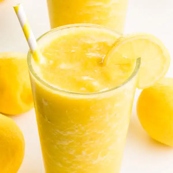 A closeup of a lemon smoothie in a glass. There is a yellow straw and a lemon slice in the glass. There are fresh lemons and another smoothie in the background.