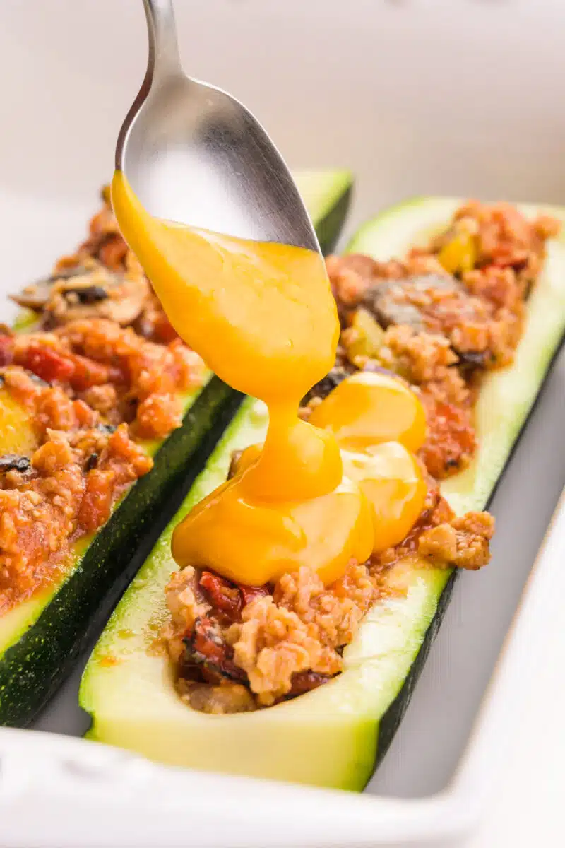 A spoon holds melted vegan cheese, drizzling it over zucchini boats.