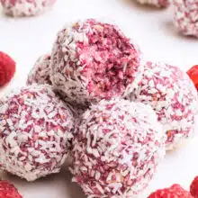 A stack of raspberry bliss balls shows the top one with a bite taken out. Fresh raspberries are scattered around the stack.