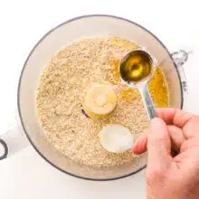 A hand pours agave nectar from a measuring spoon into a food processor bowl with other ingredients, including a scoop of coconut oil.