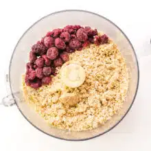 Frozen raspberries are in a food processor along with a mixture of oats and other ingredients that have been ground to a fine texture.
