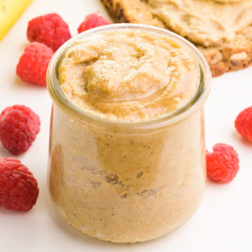 A glass jar of tiger nut butter has raspberries around it. There is a slice of toast and a banana in the background.