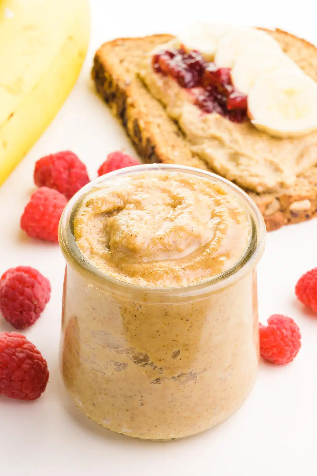 A small jar of tigernut butter has raspberries and a banana around it. There is a piece of toast with banana slices and jelly in the background.