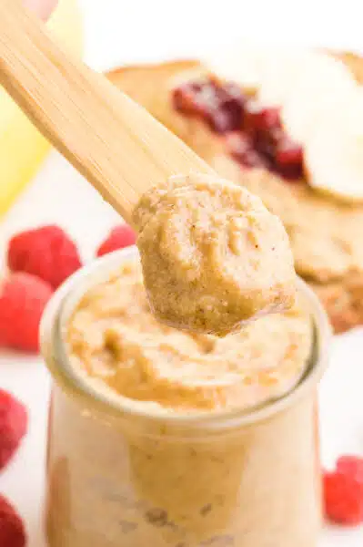 A wooden knife holds a bit of tiger nut butter on top. It hovers over a jar with more of the butter. There is fresh fruit and a slice of toast in the background.