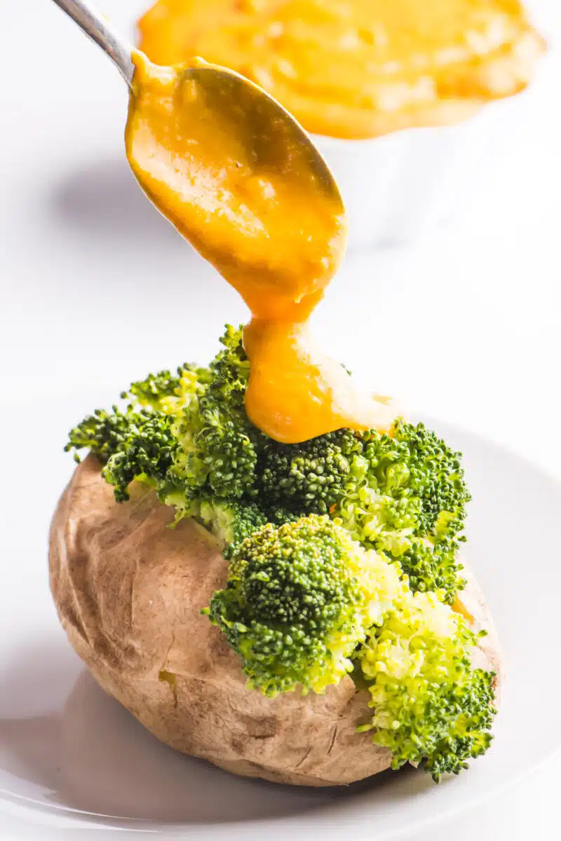 A spoon is drizzling vegan cheese sauce over a baked potato with steamed broccoli. A bowl of more cheese sauce is sitting in the background.