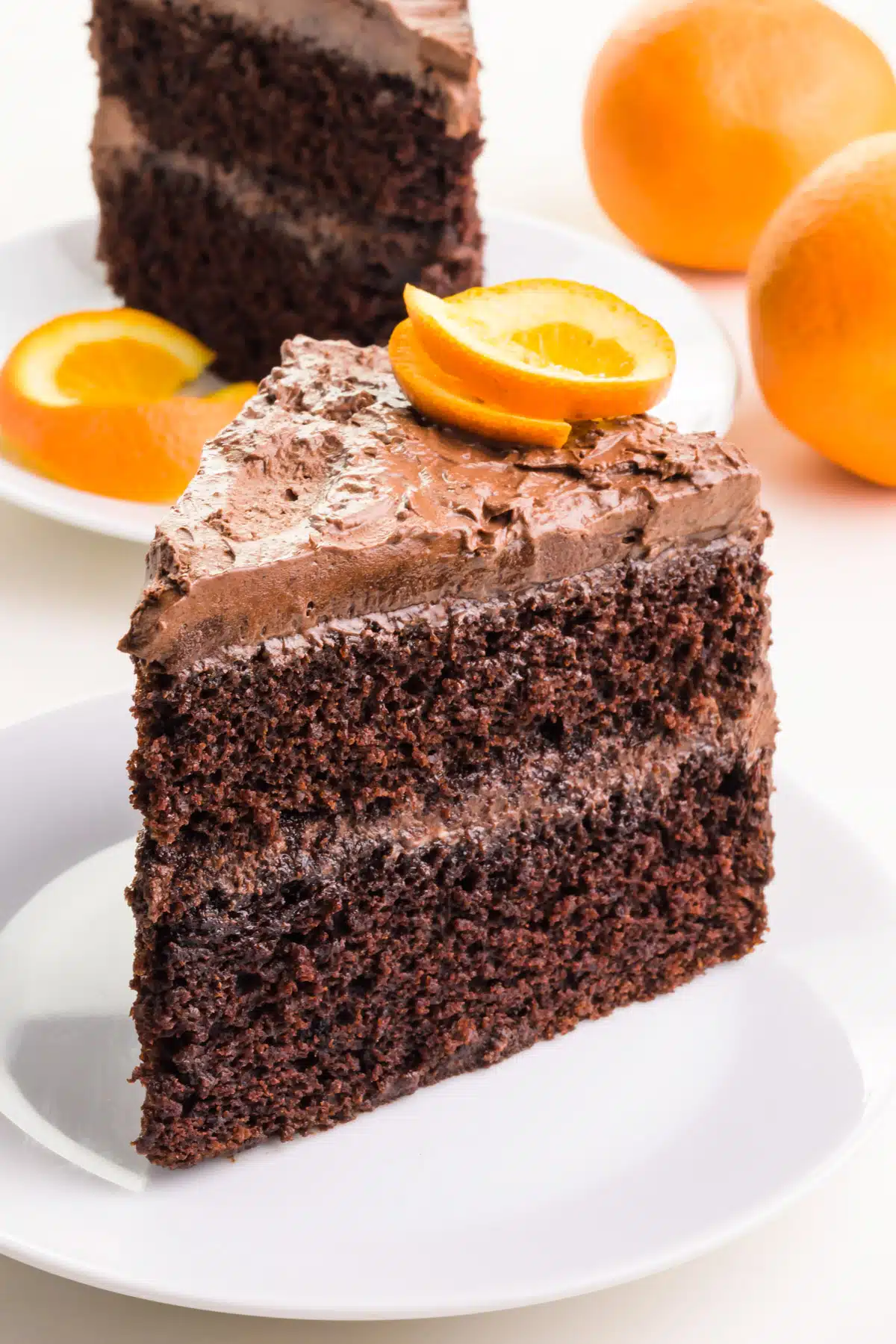 A slice of layered chocolate cake has an orange slice on top. Another slice in the background has an orange slice on the plate. There are 2 fresh oranges in the background.