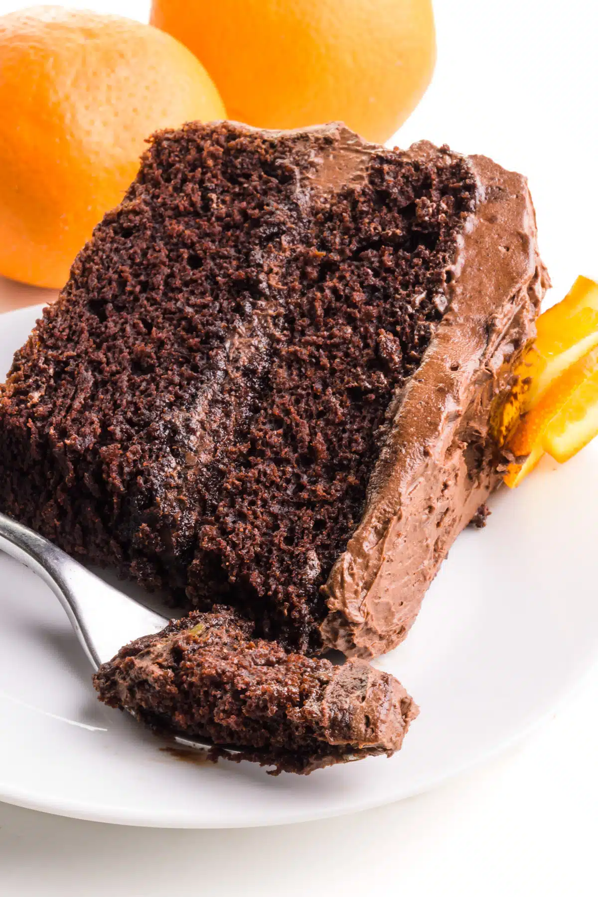 A slice of vegan chocolate orange cake is on its side on a plate. There is a fork with a bite in front of the cake. There are two oranges in the background.