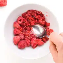 A hand holds a spoon, mashing raspberries in a bowl. There are fresh raspberries beside the bowl.