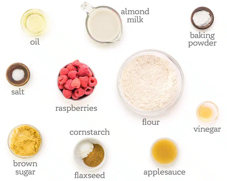 Ingredients for muffins are on a table. The text besides them reads, baking powder, vinegar, applesauce, flour, flaxseed, cornstarch, brown sugar, raspberries, salt, oil, and almond milk.