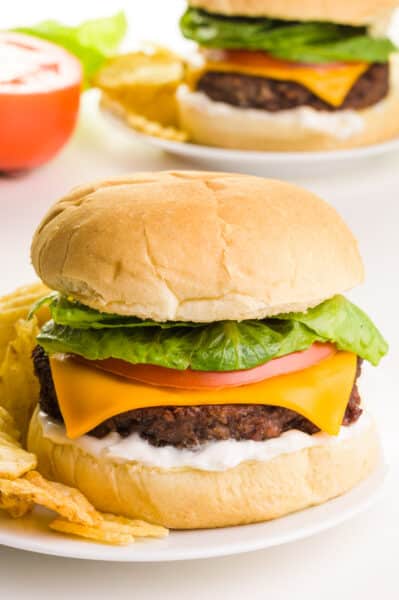 A cheeseburger is on a bun with other toppings, such as mayo, tomato, and greens. It sits on a plate with potato chips. There is another burger in the background along with a sliced tomato.