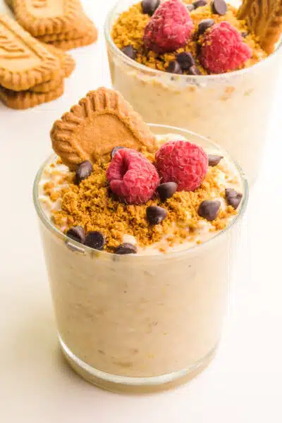 A glass jar of overnight oats has a Biscoff cookie on top with raspberries and chocolate chips. There is another jar of the oats in the background sitting beside more cookies.