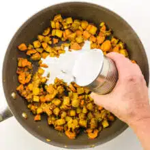 Coconut milk is being poured into a skillet with other ingredients like chopped onions and potatoes.