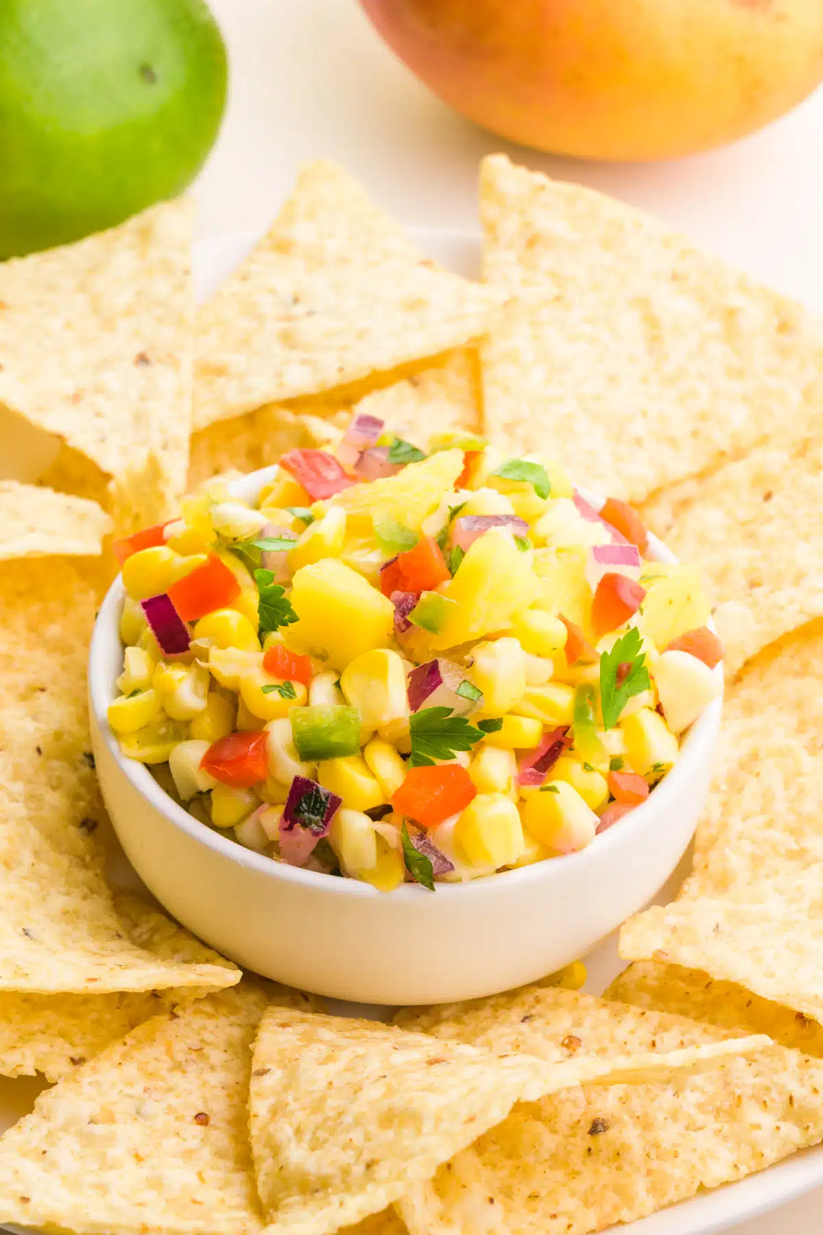 A bowl of corn mango salsa sits on a plate with tortilla chips. There is a lime and a mango in the background.