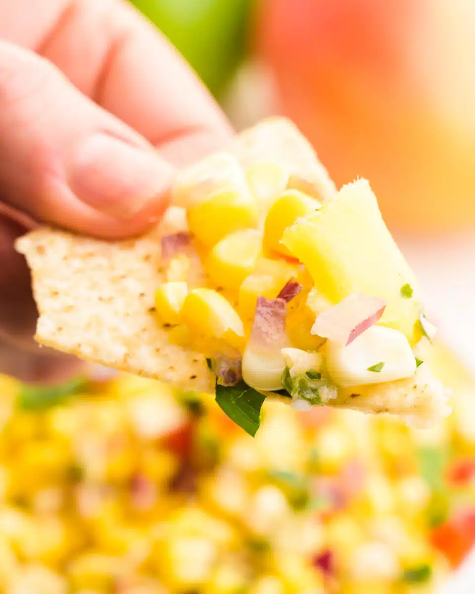 A hand holds a tortilla chip with mango corn salsa on it. The rest of the salsa is visible in the background.