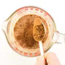 A measuring spoon of cocoa powder is being poured into a pyrex bowl with plant-based milk.