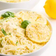 A bowl of pasta has lemon slices on top and fresh basil. There is another bowl in the background and a fresh lemon.