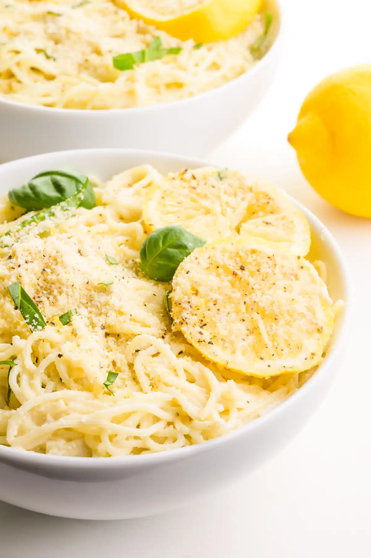 A bowl of pasta has lemon slices on top and fresh basil. There is another bowl in the background and a fresh lemon.
