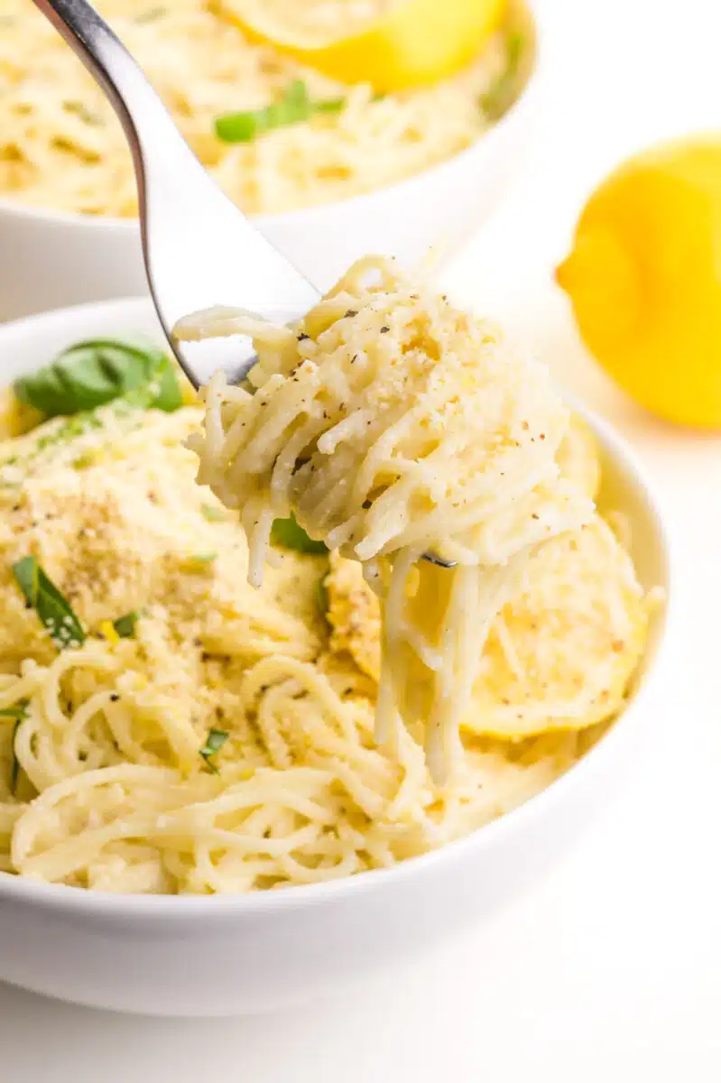 A fork holds a bite of pasta hovering over a bowl of pasta with lemons and basil on top. There is another bowl of pasta and a fresh lemon visible in the background.