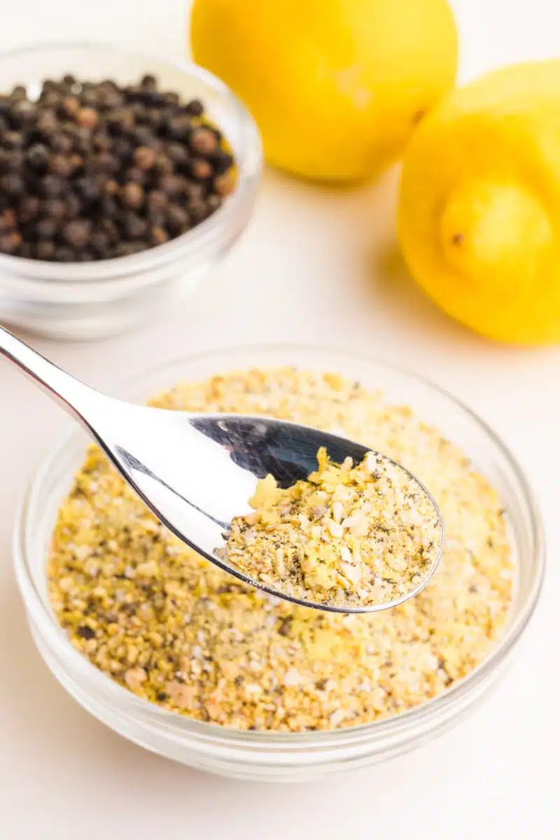 A spoon holds lemon garlic seasoning hovering over a bowl with more of the mixture. There is a bowl of peppercorns in the background along with a couple of fresh lemons.