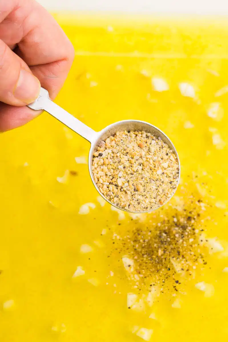 A hand holds a measuring spoon of lemon pepper seasoning, pouring it into a bowl to make a marinade.