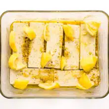 Pieces of tofu are marinading in a shallow glass dish. There are pieces of lemon scattered on top of the tofu.