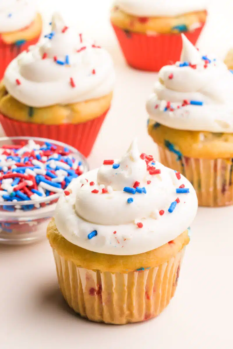A Fourth of July cupcake has white frosting on top and colorful sprinkles. There are sprinkles in a bowl behind it and more cupcakes.