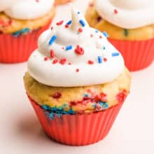 A cupcake in a red silicone cupcake liner has vanilla frosting on top. Both the cupcakes and the frosting are full of red, white, and blue sprinkles. There are more cupcakes in the background.