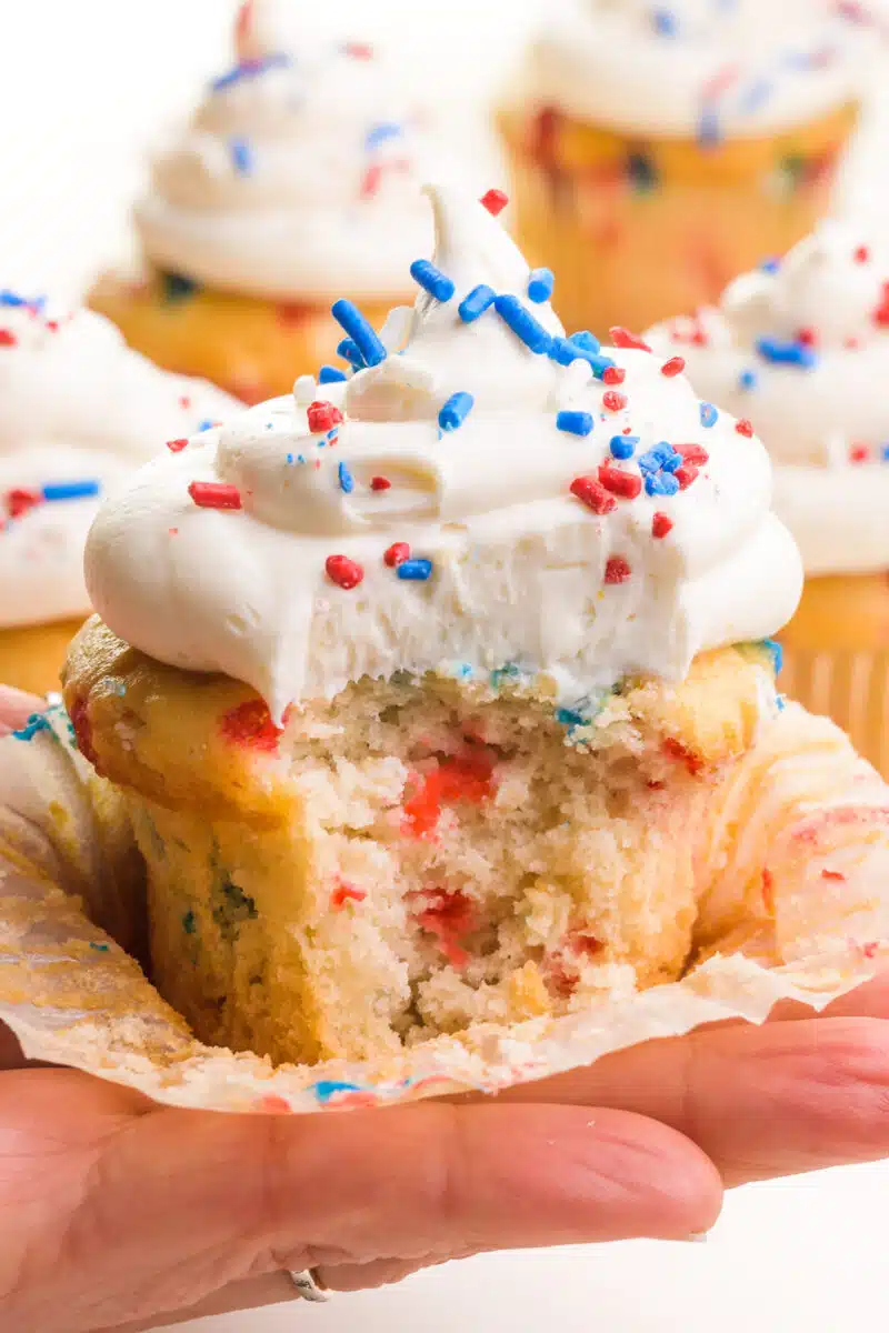 A hand holds a red, white, and blue cupcake with a bite taken out. It sits in an open cupcake liner. There is frosting on top with sprinkles. There are more cupcakes in the background.