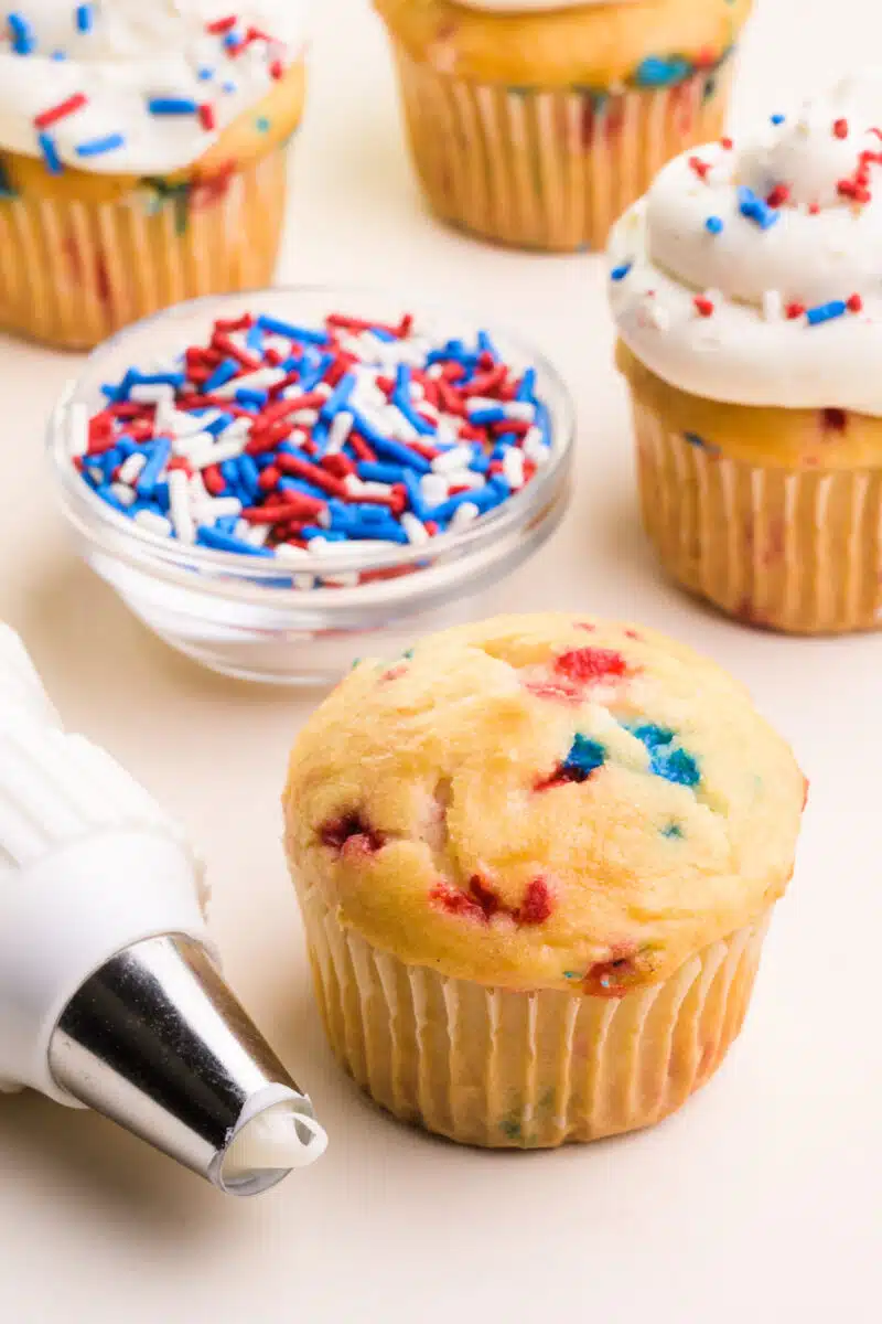 A plain cupcake has red and blue sprinkles visible in the top. It sits beside a pastry bag with vanilla frosting. Behind it is a bowl of red, white, and blue sprinkles and more cupcakes that are frosted.