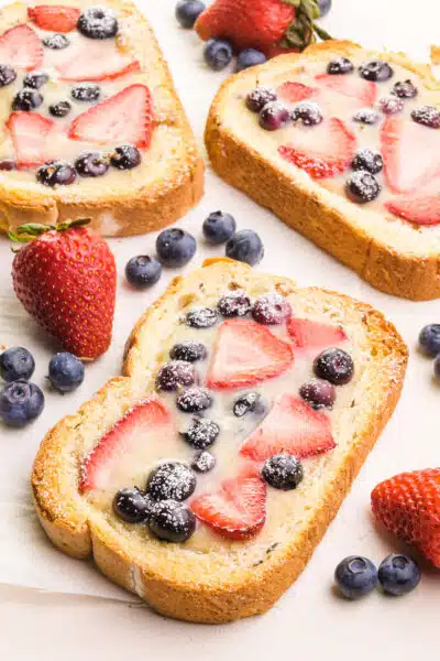Looking down on several slices of vegan custard toast with fruit on top. There are fresh strawberries and blueberries beside each slice.
