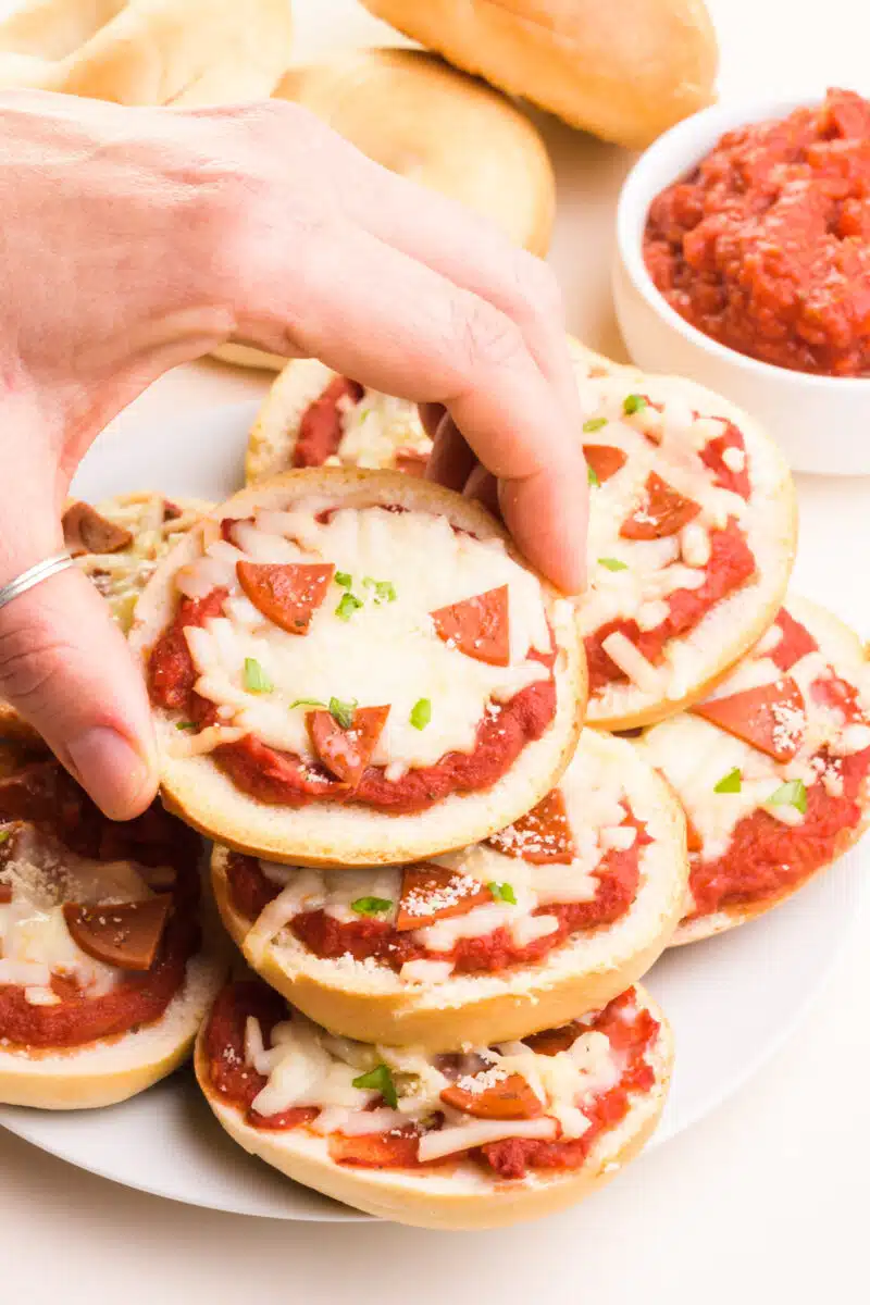 A hand reaches down to grab a vegan pizza bagel from a stack on a plate. There is a bowl of marinara in the background.