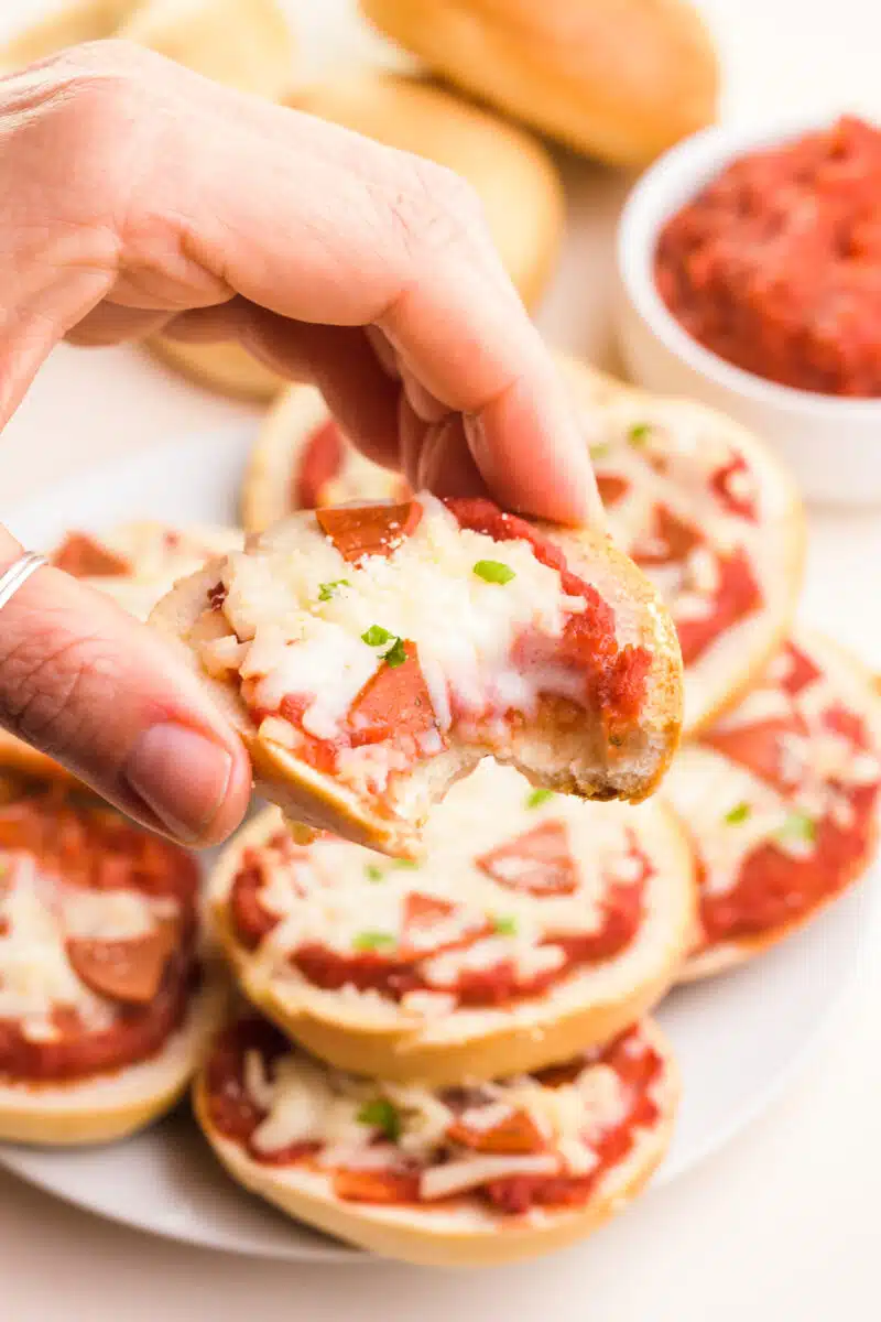 A hand holds a vegan pizza bagel with a bite taken out, revealing melted vegan cheese. There is a plate with more of the bagels below it and a bowl of red sauce in the background.