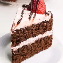A slice of chocolate layer cake has strawberry frosting with a fresh strawberry and chocolate sauce on top.