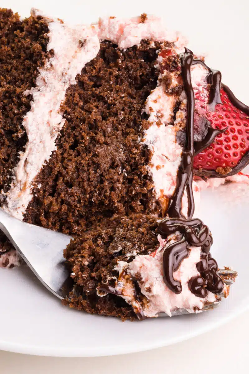 A slice of vegan chocolate strawberry cake lays on its side on a plate. A bite on a fork sits in front of the cake.