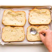 Four slices of bread are in a baking pan. A hand holds a spoon, pressing in the center to create a well in each slice of bread.