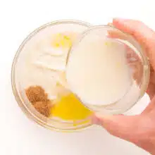 A hand holds a bowl with a "flour egg", pouring it into another bowl with yogurt and spices.