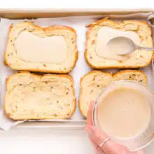 A hand holds a bowl of custard, and is using a spoon to distribute into four slices of bread in a baking pan.